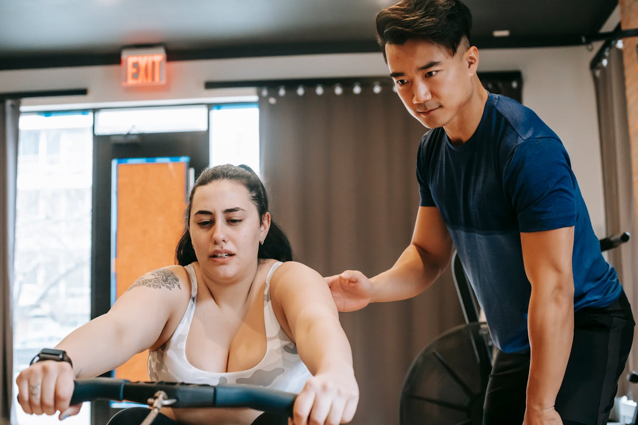 About Young handsome ethnic experienced trainer supporting plus size woman working out on rowing machine in gym